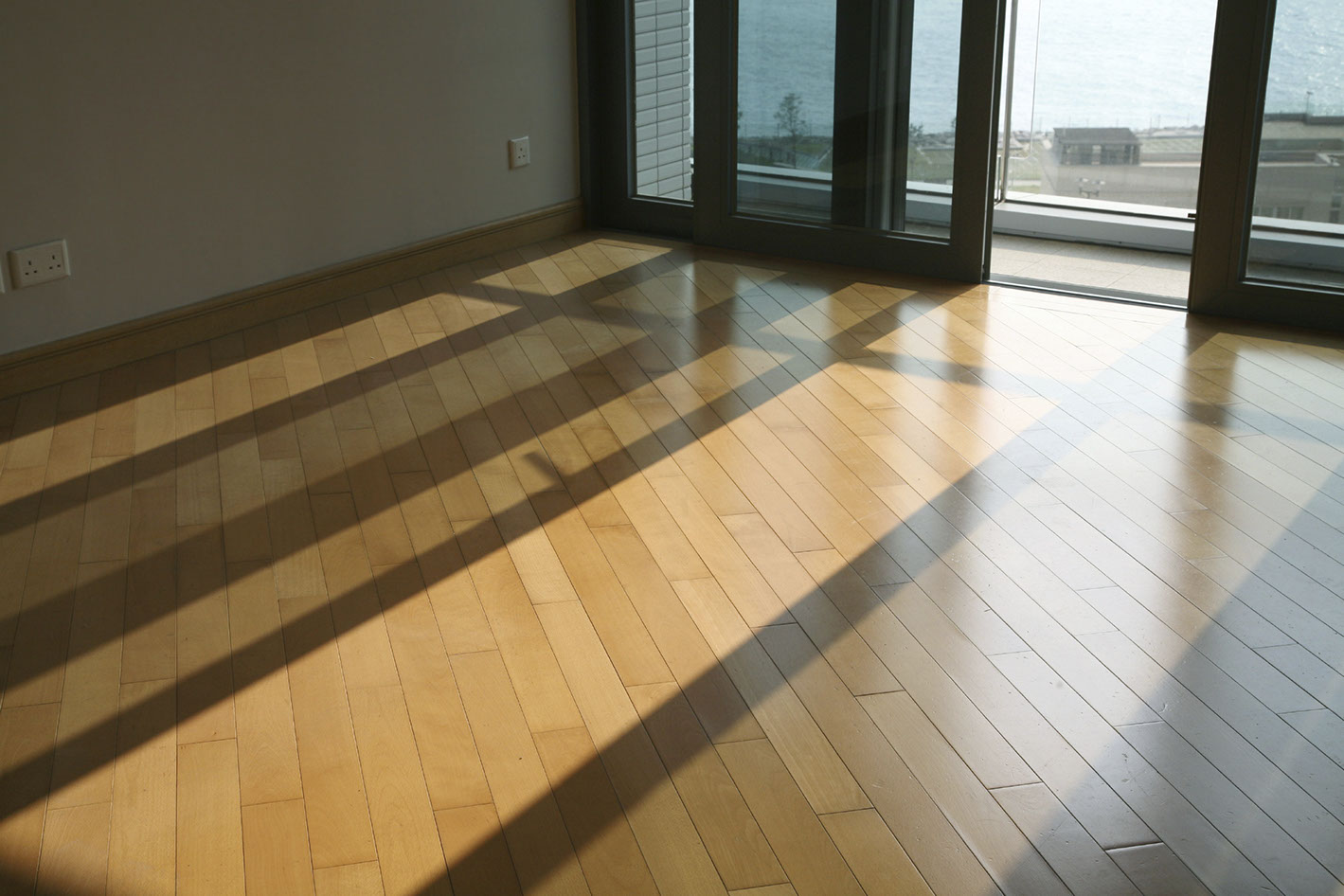 Détail d'une chambre avec parquet. En fond, une baie vitrée est à moitié ouverte. La lumière au sol est filtrée, sauf sur la partie ouverte. 